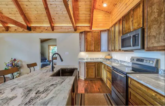 A rustic cabin style kitchen featuring dramatic grey and white veined granite countertops on a large island with an under-mount sink, paired with warm natural wood cabinetry and a vaulted wood-plank ceiling.